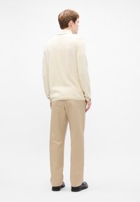 Beige long-sleeve shirt with a soft texture and ribbed cuffs, paired with light beige trousers, worn with black shoes. Simple, neutral design.
