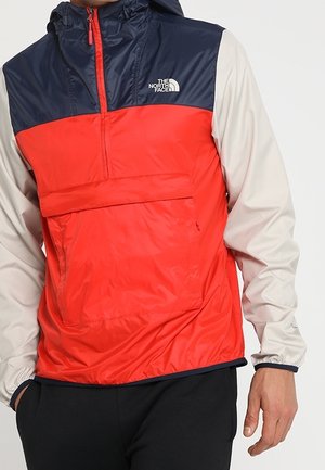 Veste coupe-vent rouge et marine avec un design à demi-zip, grande poche avant, manches élastiques et capuche. Fabriquée en matériau léger et lisse.