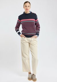 Maglione a righe in blu navy, rosa e bianco con colletto alto e dettagli con bottoni; abbinato a pantaloni larghi color crema e sneakers casual.