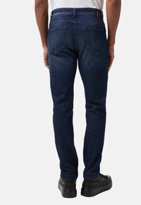 Dunkelblaue Jeans aus Denim mit geradem Schnitt, sichtbaren Gesäßtaschen und einem Lederpatch am Bund. Getragen mit schwarzen Schnürschuhen.