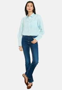 Naine seisab käed lõdvalt langenud, seljas hele sinise ja valge triibuline nööpidega särk, tumedad sinised teksad ja pruunid baleriinakingad.