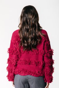 Pull en maille rouge avec des détails en frange texturés sur les manches et le corps, associé à un pantalon rayé gris. Vue de dos.