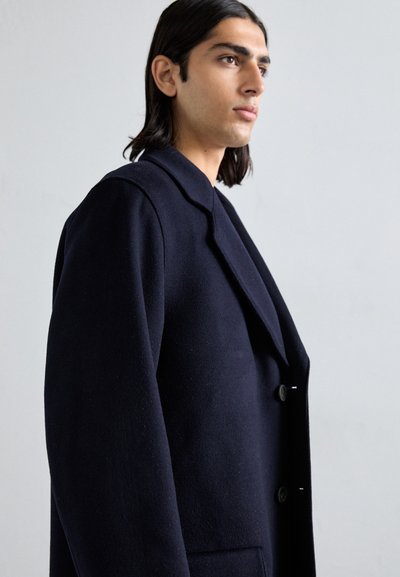 Cappotto in misto lana blu navy con colletto a soffietto, chiusura doppiopetto e tasche laterali. Superficie testurizzata con un fit leggermente oversize.