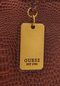 Sac en cuir marron texturé avec une étiquette dorée portant l'inscription "GUESS EST 1981" gravée. L'étiquette est fixée avec un anneau doré circulaire.