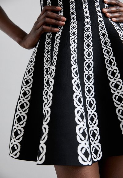 Rochie tricotată neagră cu modele verticale din fir alb, care seamănă cu noduri complicate, având un design cu fustă evazată și un material texturat.