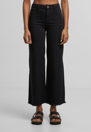 Person, die schwarze High-Waist-Jeans mit weitem Bein, ausgefransten Säumen und schwarzen Slip-On-Sandalen trägt, vor einem einfarbigen grauen Hintergrund stehend.