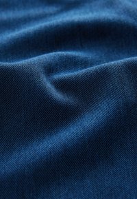 Blauwe denimstof met een gladde textuur, voorzien van subtiele diagonale ribbels en zachte plooien, die het licht in verschillende tinten vangt.