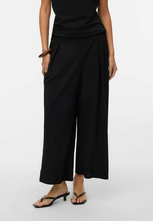 Kvinne som har på seg svarte vide bukser, svart ermeløs topp, brun armbånd og svarte sandaler med hæl på hvit bakgrunn.