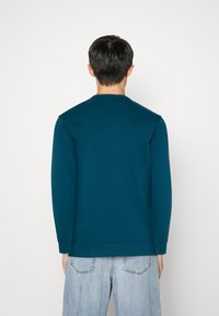 Homme avec des cheveux courts et foncés portant une chemise à manches longues teal et un jean bleu clair, faisant face à un fond blanc uni.