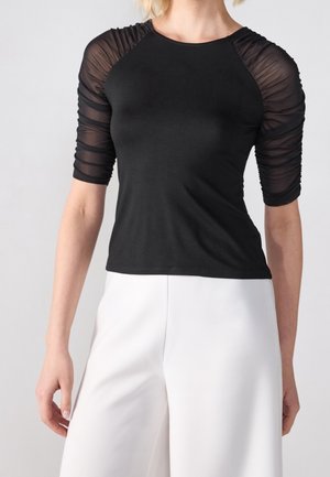 Czarna dopasowana bluzka z przezroczystymi, marszczonymi rękawami. Okrągły dekolt i gładkie, jednolite body. W zestawieniu z białymi, szerokimi spodniami dla kontrastu.