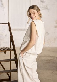 Femme en gilet blanc sans manches et pantalon assorti, debout les mains dans les poches près d'une échelle en bois dans une pièce spacieuse et lumineuse.