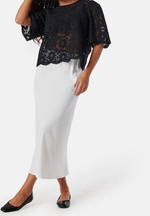 Top de encaje negro con mangas cortas que cubre una falda blanca ajustada, con una textura suave y un diseño hasta el suelo, complementado con zapatos planos negros.