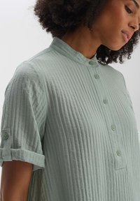 Hellgrünes, strukturiertes Hemd mit Stehkragen, drei Knöpfen und aufgerollten Ärmeln. Der Stoff weist ein feines geripptes Muster auf.