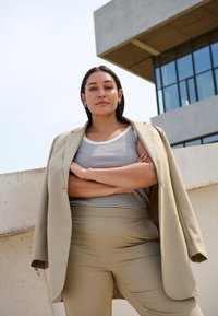 Beige blazér přes pruhované tílko, doplněný o světlé béžové úzké kalhoty. Žena stojí se zkříženýma rukama před betonovým pozadím.