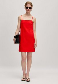 Rotes ärmelloses Kleid mit dünnen Trägern, geradem Schnitt und glatter Textur. Kombiniert mit einer schwarzen Clutch und minimalistischen schwarzen Sandalen.