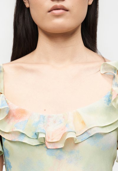 Νεαρή γυναίκα με μακριά σκούρα μαλλιά που φοράει ένα παστέλ λουλουδάτο φόρεμα με γυμνούς ώμους, διακοσμημένο με βολάν στο ντεκολτέ και ακάλυπτα κολάρα.