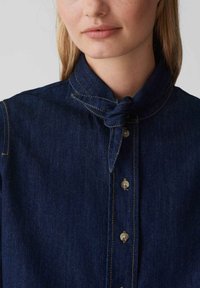 Camicia di jeans scuri con colletto annodato, dotata di cuciture a contrasto chiare e chiusura frontale con bottoni piccoli e rotondi.