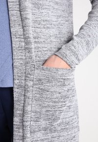Cardigan grigio lavorato a maglia con un motivo testurizzato, dotato di maniche lunghe e una tasca laterale. Il tessuto appare morbido e leggero.