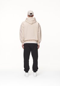 Felpa beige con un ampio cappuccio, polsini e vita a costine, abbinata a pantaloni jogger neri. Sneakers bianche completano l'outfit.