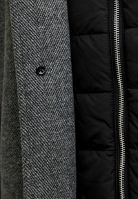 Grijs wollen jasje met een houndstooth patroon en een zwarte gewatteerde voering, voorzien van een zwarte knop en een zilveren rits. Textuurcontrasten tussen de stoffen.