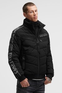 Mann trägt eine schwarze gesteppte Jacke mit Reißverschlüssen und dem Schriftzug "CAMP DAVID" auf Brust und Ärmeln, steht vor einem einfarbigen Hintergrund.