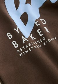Hnědá mikina s velkým světle modrým logem a bílým textem "BY BAKER ESTABLISHED NINETEEN EIGHTY" s hladkou texturou.
