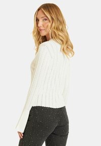 Suéter blanco de punto con textura acanalada y mangas acampanadas, combinado con jeans de mezclilla oscura que presentan acentos de tachuelas plateadas.