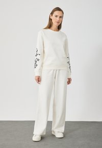 Sudadera blanca con bordado floral negro en las mangas, combinada con pantalones blancos holgados a juego, exhibida sobre un fondo neutro.