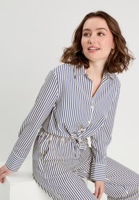 Ensemble blouse et pantalon rayé en bleu et crème, avec un devant à boutons, un col et une taille à nouer. Tissu lisse avec des manches longues et des poignets.