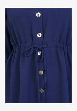 Navyblaue Knopfleiste mit Zugband in der Taille, ausgestattet mit großen goldfarbenen Knöpfen und einem strukturierten, leichten Stoff.