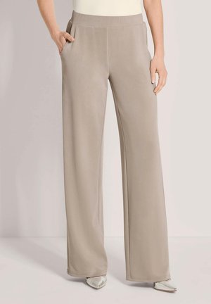 Beige gerade Hose mit elastischem Bund, seitlichen Taschen, getragen von einer Person mit einer Hand in der Tasche und silbernen spitzen Schuhen.