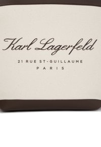 Kangas laukku, jossa on kerma sävyinen runko ja ruskeat nahkakoristeet. Kyljessä on mustaa tekstiä, jossa lukee "Karl Lagerfeld" ja "21 RUE ST-GUILLAUME, PARIS."