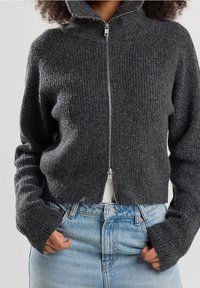 Suéter de punto acanalado gris oscuro con cremallera y cuello alto, silueta ajustada y mangas largas, combinado con jeans de denim claro.