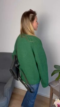 Suéter verde de malha com um corte folgado, usado com jeans de ganga azuis. Uma mala de mensageiro preta pende sobre o ombro. Piso de madeira clara.