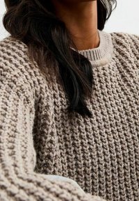 Gruba sweter w jasnym taupe z teksturowanym splotem. Posiada prążkowany okrągły dekolt i opuszczone ramiona, co zapewnia luźny krój.
