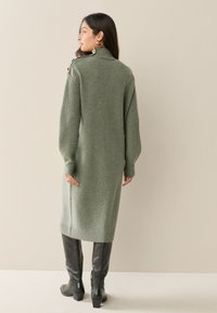 Abito verde lavorato a maglia con collo alto, maniche a coste e piccoli dettagli con bottoni. Abbinato a stivali neri al ginocchio. Colore uniforme, texture morbida.