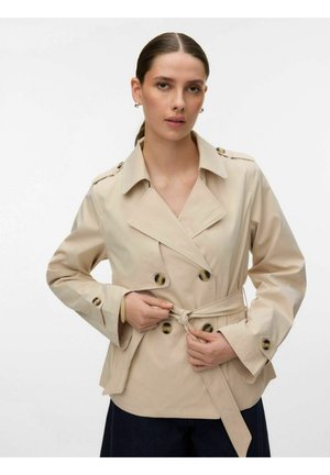 Kobieta o brązowych włosach związanych do tyłu, ubrana w beżowy dwurzędowy trencz, zapinająca pasek, stojąca na jednolitym białym tle.