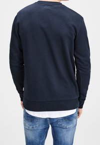 Uomo che indossa una felpa a maniche lunghe blu navy sopra una camicia bianca, con jeans blu, visto di spalle su uno sfondo neutro.