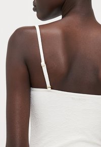 Biely bikini top s textúrovaným listovým vzorom, s tenkými ramienkami a zlatým klipom na nastavenie pre lepšie prispôsobenie. Nápis značky "BILLABONG" je viditeľný.