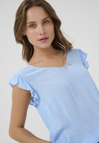 Haut bleu clair avec des manches courtes à volants, un décolleté en V et un tissu texturé. Présente une poche discrète d'un côté.
