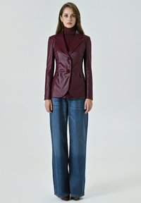 Donna in piedi con un blazer di pelle bordeaux sopra un dolcevita coordinato e jeans a gamba larga blu, su uno sfondo neutro.