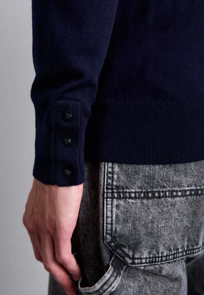 Close-up of a hand next to a navy blue sweater cuff with three black buttons and gray denim jeans pocket.