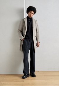 Abrigo de mezcla de lana beige sobre un polo negro, combinado con pantalones de rayas azul marino y zapatos negros gruesos, con un fondo claro.