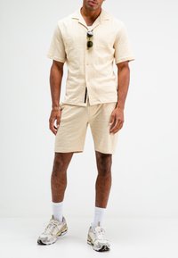 Beige textuur knoopshirt en bijpassende shorts. Draagt het met getinte zonnebrillen en gebloemde sneakers. Witte crew-sokken maken de look af.