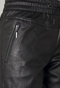 Svarta joggers i konstläder med elastiskt midjeband, dragsko och sidoficka med dragkedja. Ytan har en något texturerad finish.