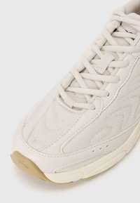 Beige mesh and synthetic sneaker with textured overlays, padded tongue, and flat laces; rubber sole with slight tread for traction.