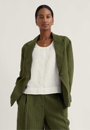 Žena v olivovo zelenom obleku s jemnými pruhmi, voľné nohavice a sako cez bielu blúzku, doplnené zlatými náušnicami a náhrdelníkom.