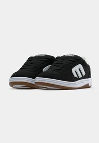 Μαύρα χαμηλά αθλητικά παπούτσια με λευκές σόλες, μαύρους κορδόνια, λευκή επένδυση στο εσωτερικό και λευκό λογότυπο "M" στο πλάι από την Etnies.