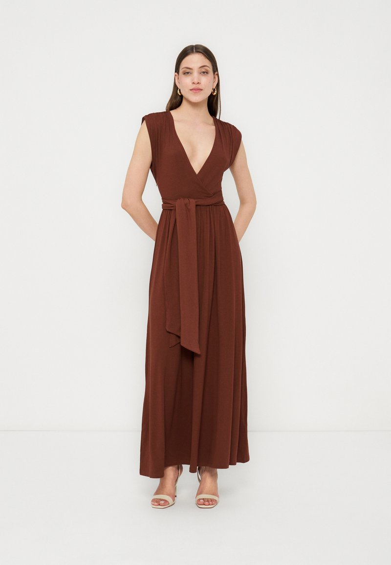 Femme debout portant une robe portefeuille sans manches marron avec un col en V profond et une taille nouée, associée à des escarpins beige à bouts ouverts, sur un fond uni.