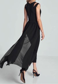 Frau in einem ärmellosen schwarzen Kleid mit einem durchsichtigen, fließenden Überrock über enganliegenden schwarzen Hosen und schwarzen High Heels, die vor einem schlichten Hintergrund davongeht.